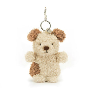 Jellycat Little Pup Bag Charm. The small, plush puppy has fluffy nougat fur with brown patches, floppy ears, and a playful grin. It is attached to a silver clasp, ready to be clipped onto a bag or backpack