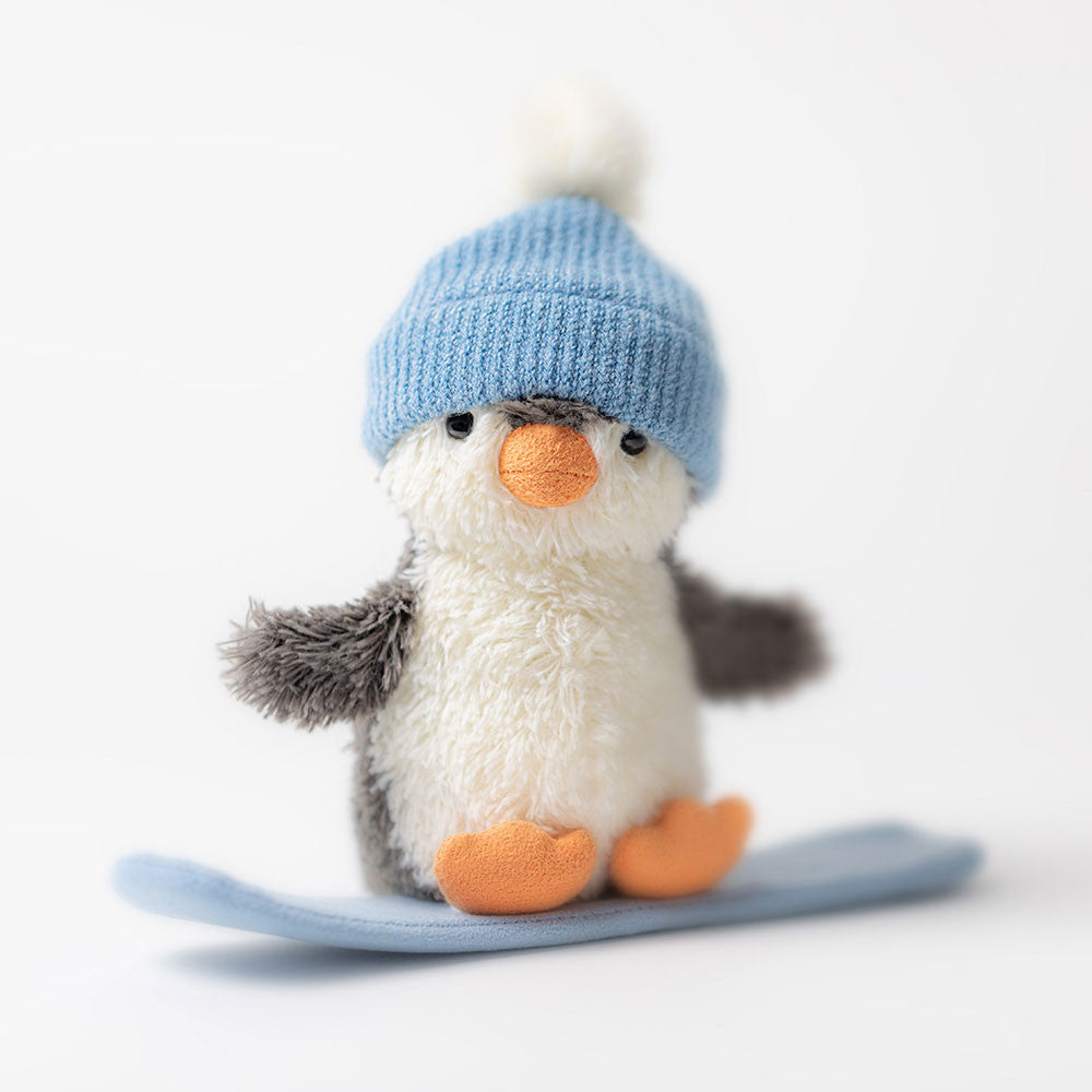 Close-up of Jellycat Peanut Penguin with blue knitted hat and orange beak on snowboard.