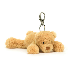 Angled View: Golden cuddles on the go! The Jellycat Smudge Bear Bag Charm tilts forward, showcasing its warm golden fur with a sweet embroidered face and a cute, bobble tail. Holdable paws and a secure silver claw clip let Smudge add a touch of whimsy to any bag.