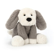 Angled view: Jellycat Smudge Puppy sitting up,  features soft fur, a sleepy expression, floppy ears, and a wagging tail.