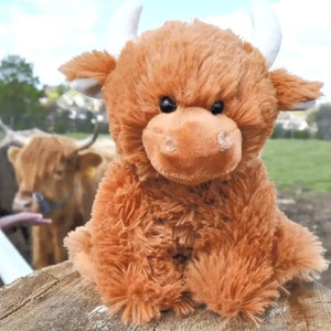 The children's soft toy highland cow sitting on a fence outside with a real highland cow in the background. 