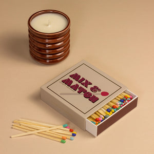 Stylish lifestyle shot featuring the Kiln Mix & Match matchbox next to a ribbed brown ceramic candle, with colourful matches scattered across a beige background.