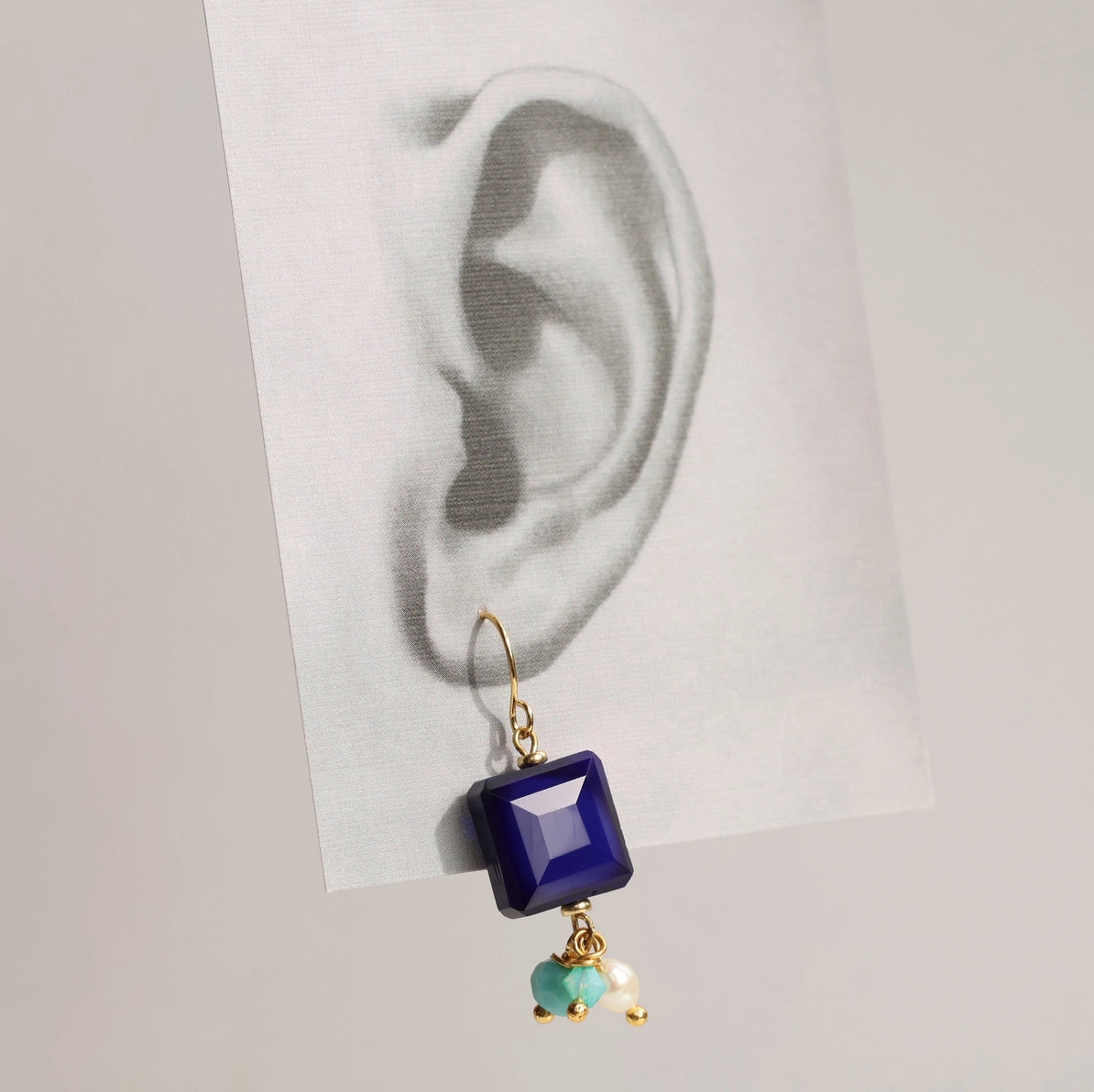 Single Lapis Lazuli blue charm earring hanging on a black-and-white ear illustration, showcasing geometric navy glass stone and trio of turquoise and pearl charms.