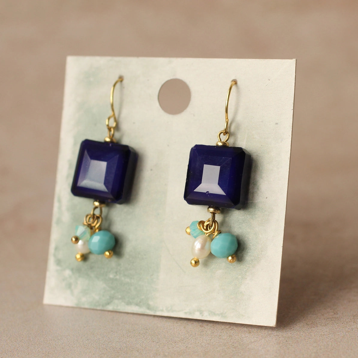 Pair of Lapis Lazuli blue charm earrings displayed on a textured green and white backing card, highlighting the gold-tone hooks and colourful bead drops.