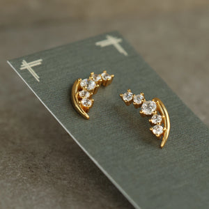 Angled shot of gold-plated sterling silver lily of the valley stud earrings on a green display card, showing the curved stem and sparkling crystal detailing.