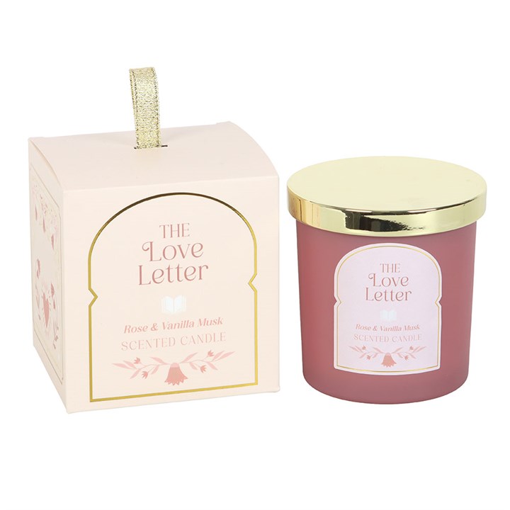 The Love Letter Rose & Vanilla Musk Candle in pink jar with gold lid displayed next to decorative illustrated gift box with gold ribbon handle.