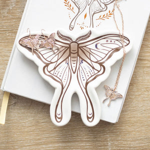 Close-up of Luna Moth ceramic trinket dish holding gold jewellery on a book, showing detailed moth design in brown and blush tones.
