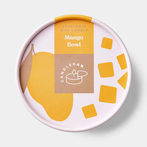 Mango-scented tin candle placed on a white book next to a bed, with open lid revealing realistic cubed wax design, creating a warm, summery ambiance in a cosy bedroom setting.