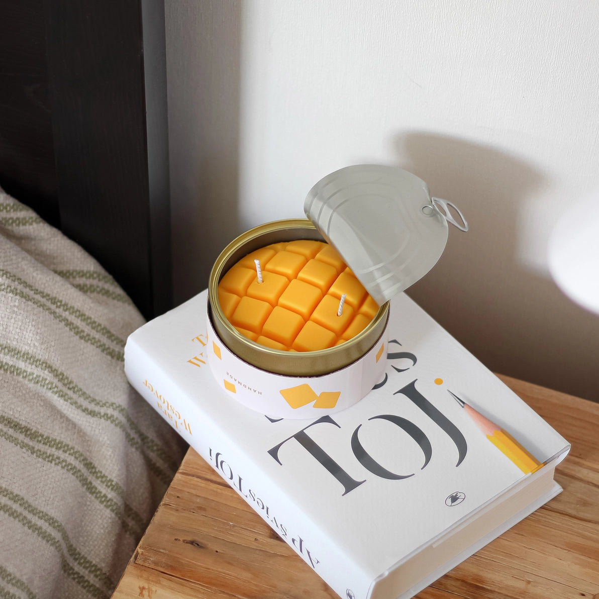Mango-scented tin candle placed on a white book next to a bed, with open lid revealing realistic cubed wax design, creating a warm, summery ambiance in a cosy bedroom setting.