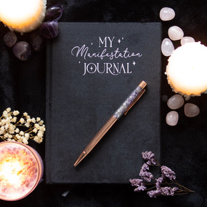 Black velvet manifestation journal with amethyst pen on dark background surrounded by candles and crystals.