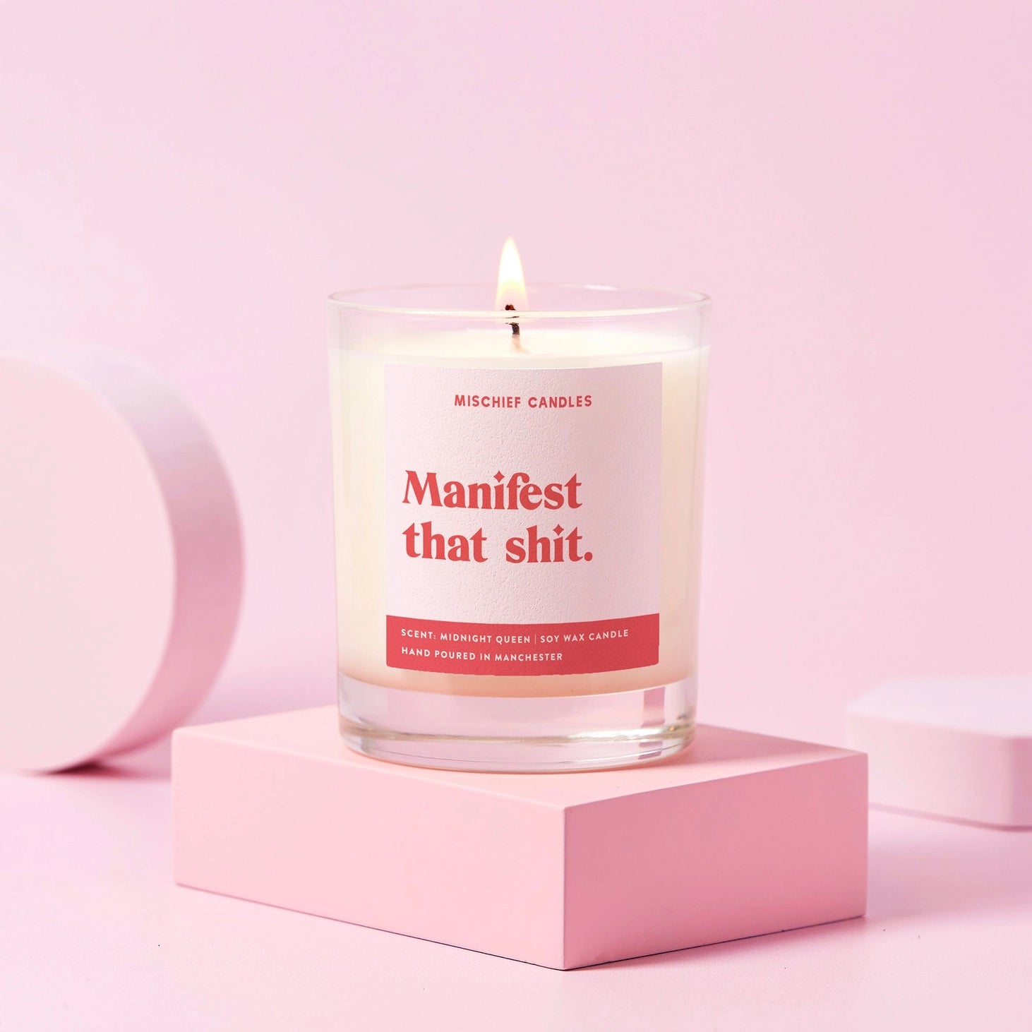 Manifest That Shit candle in glass jar with pink and red label, Midnight Queen scent soy wax candle, hand poured in Manchester, styled on pink blocks.