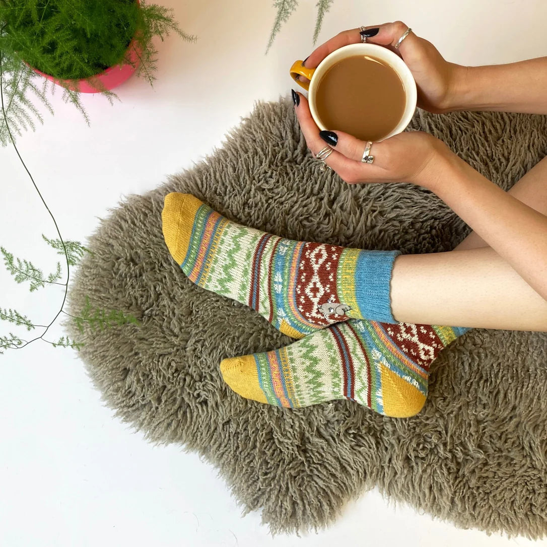 Person wearing colourful Moomin Fair Isle socks while holding a mug of tea, cosy on a shaggy rug.