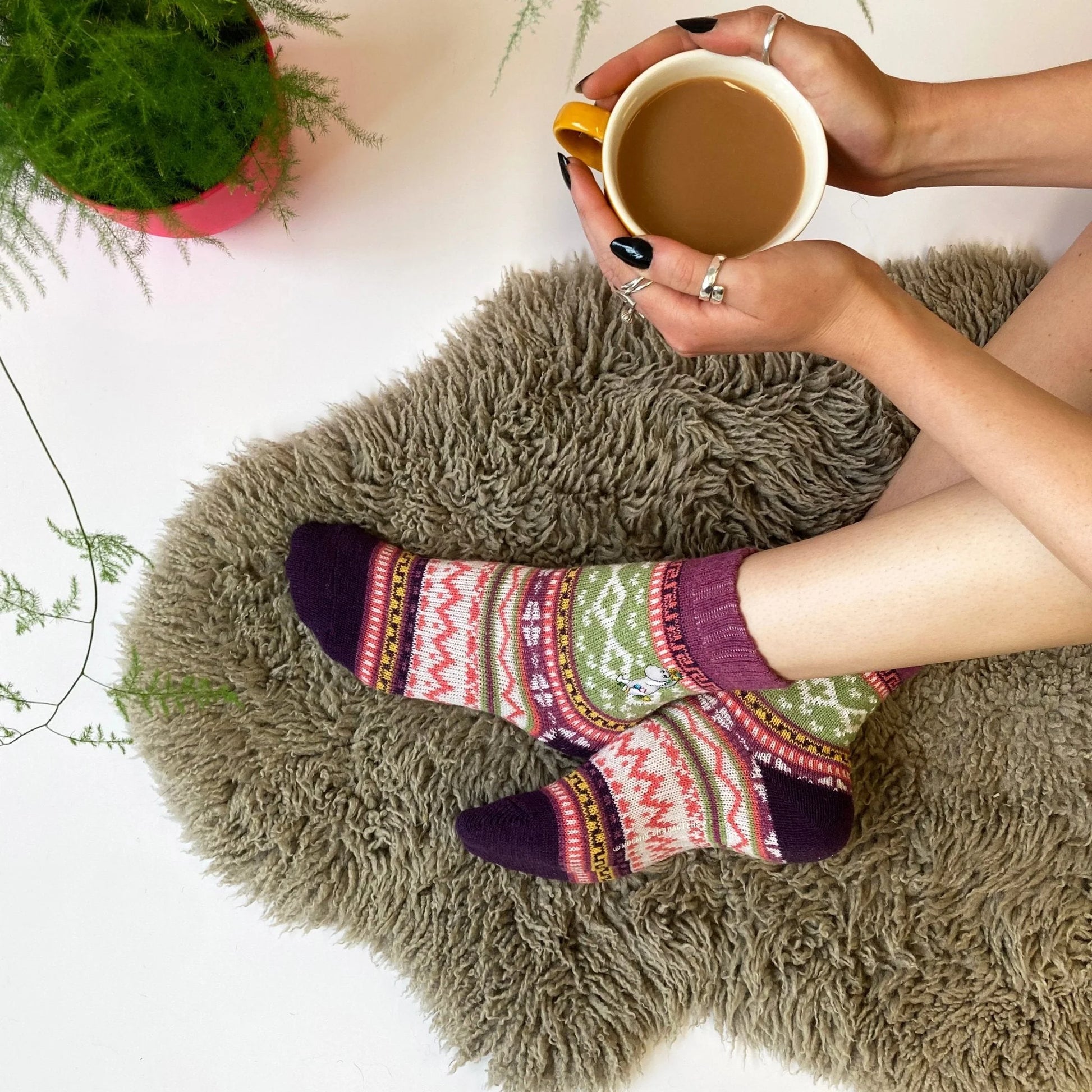 Woman wearing Moomin Fair Isle Snorkmaiden socks on cosy rug with a mug of tea.