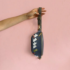 Moomin Family vegan leather bum bag in dark blue with grey strap, gold hardware, and Moomin characters print, held against pink background.
