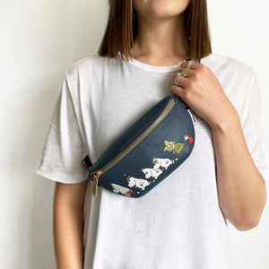Moomin Family vegan leather bum bag in dark blue with grey strap, gold hardware, and Moomin characters print, held against pink background.