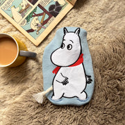 Baby blue Moomin mini hot water bottle with knit cover and applique design on fluffy rug beside comic and tea.