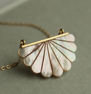 Mother of Pearl Scallop Necklace on a textured surface – A beautifully iridescent scalloped Mother of Pearl pendant with a gold-plated bar and chain, showcased against a soft green background.