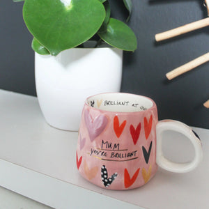Pink heart mug on a shelf near a plant – “Mum You’re Brilliant” mug styled on a modern shelf with plant.