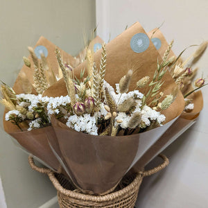 A beautifully arranged Natural Dried Flower bouquet wrapped in brown paper, displayed in a woven basket.