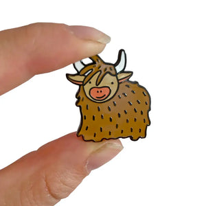 Cute enamel Highland Cow pin badge with cartoon-style design, shown being held between two fingers for scale – Scottish accessory for bags, hats, and clothes.