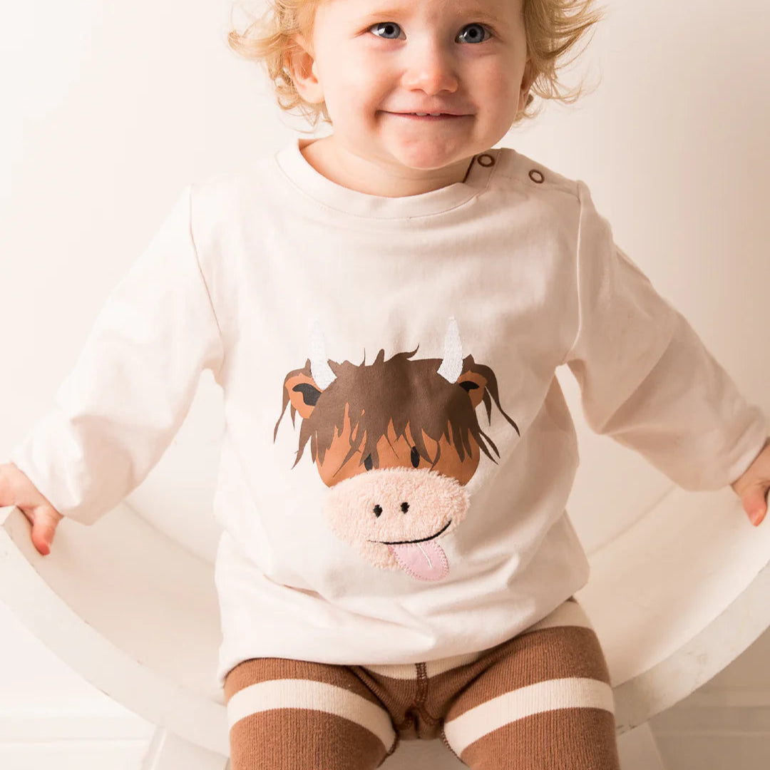 Toddler wearing Neutral Highland Cow top with soft fleece nose design, smiling while sitting on a chair.