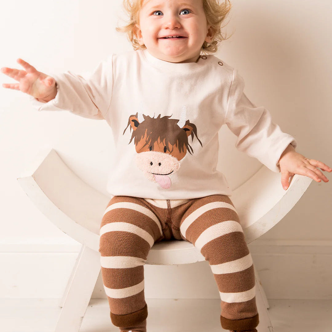 Toddler happily sitting and wearing a Neutral Highland Cow top with matching leggings, smiling at the camera.
