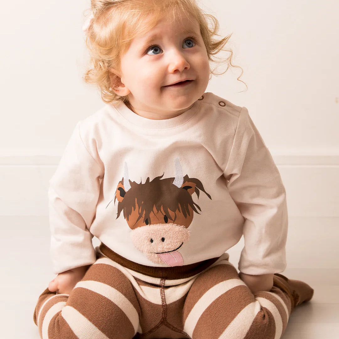Child sitting on the floor wearing Neutral Highland Cow top, looking up with an adorable expression.