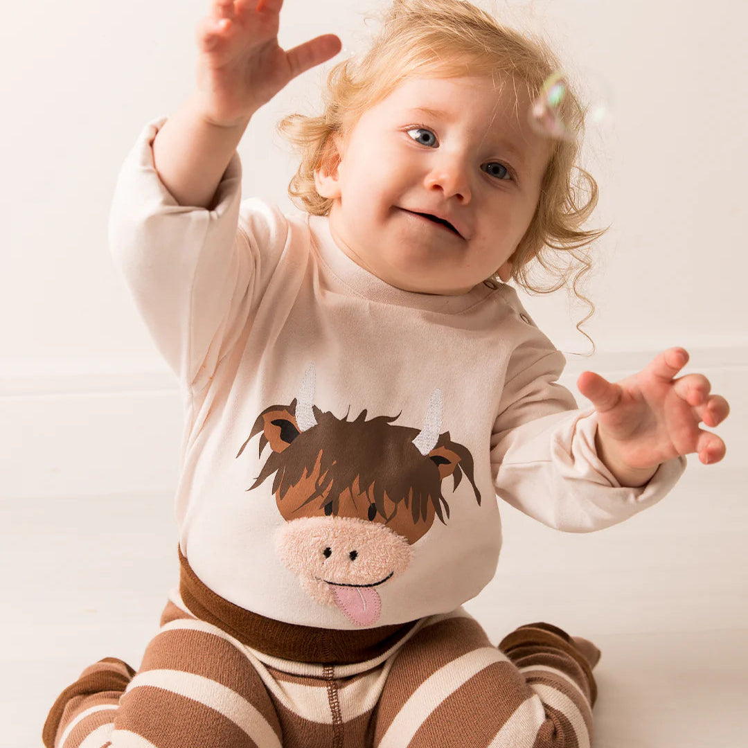 Baby reaching for bubbles while wearing a soft Neutral Highland Cow top, featuring a playful Hamish Highland Cow print.