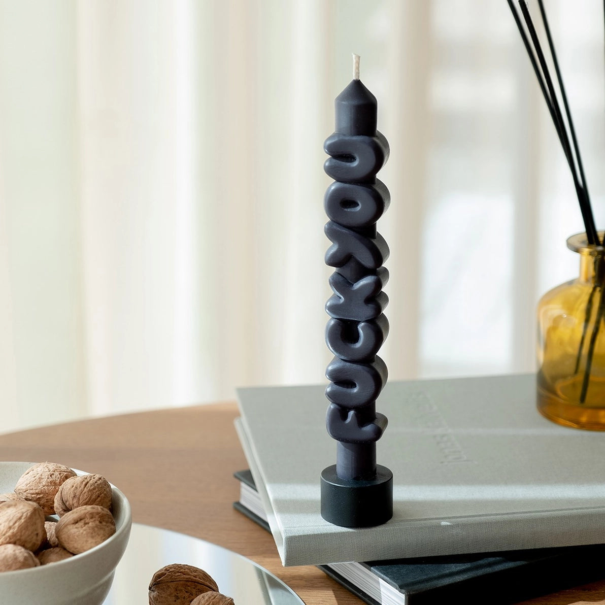 Black Nine Inch Fck You candle standing upright on stacked books next to a reed diffuser, with bold sculpted lettering and a modern interior background.