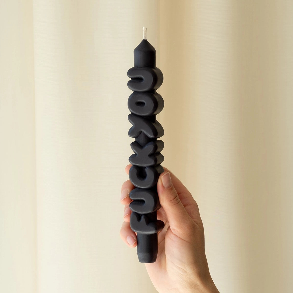 Hand holding the black Nine Inch Fck You taper candle vertically, showing sculpted block letters and matte wax finish against a cream curtain backdrop.