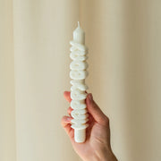Person holding a white 9-inch handmade taper candle with raised 3D “HAPPY BDAY” lettering, made from soy-paraffin wax blend, against a neutral curtain background.