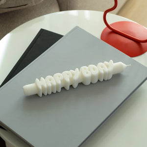 White 3D “HAPPY BDAY” taper candle resting on stacked grey books next to a red lamp, showcasing playful design and gift-ready presentation.