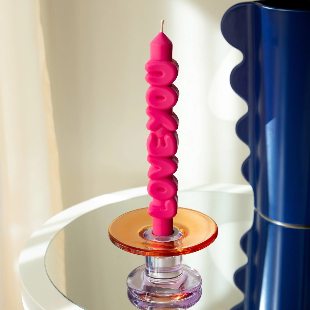Pink sculpted “LOVE YOU” candle displayed in a colourful glass candle holder on a mirrored surface, next to a wavy blue vase.