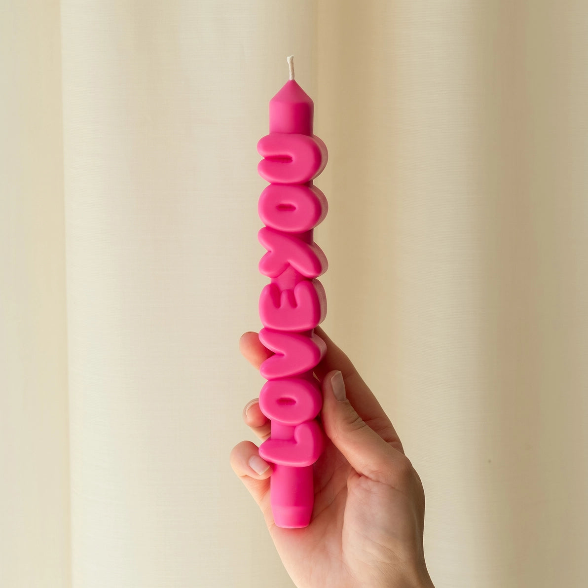 Hand holding a pink taper candle that spells “LOVE YOU” in bold, playful letters, with a soft cream curtain in the background.