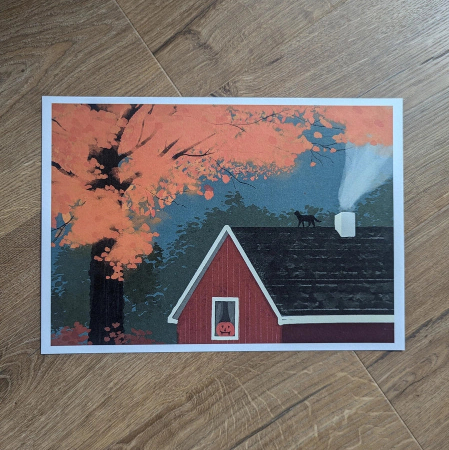 October Landscape Art Print displayed on wooden floor, showing cosy red cabin in autumn landscape illustration.