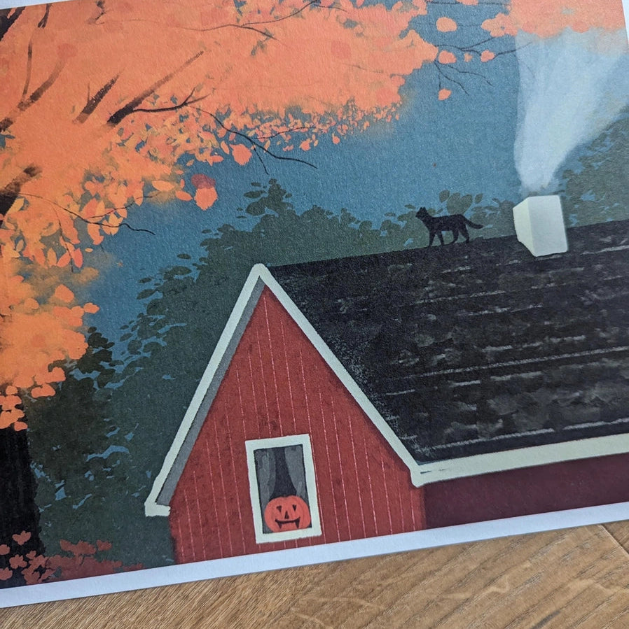 Hand holding October Landscape Art Print featuring autumn cabin, jack-o’-lantern window, and golden tree.