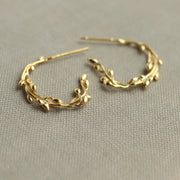 A pair of gold-plated sterling silver olive branch hoop earrings lying on a soft fabric surface.