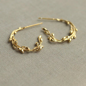 A pair of gold-plated sterling silver olive branch hoop earrings lying on a soft fabric surface.