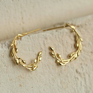 A pair of gold-plated sterling silver olive branch hoop earrings displayed on a textured stone surface.