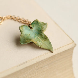 Olive Green Ivy Leaf Necklace resting on a book, highlighting its brass finish and organic design.