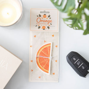 Orange slice air freshener in packaging on a white desk with candle, car key, and plant – citrus-themed car accessory.