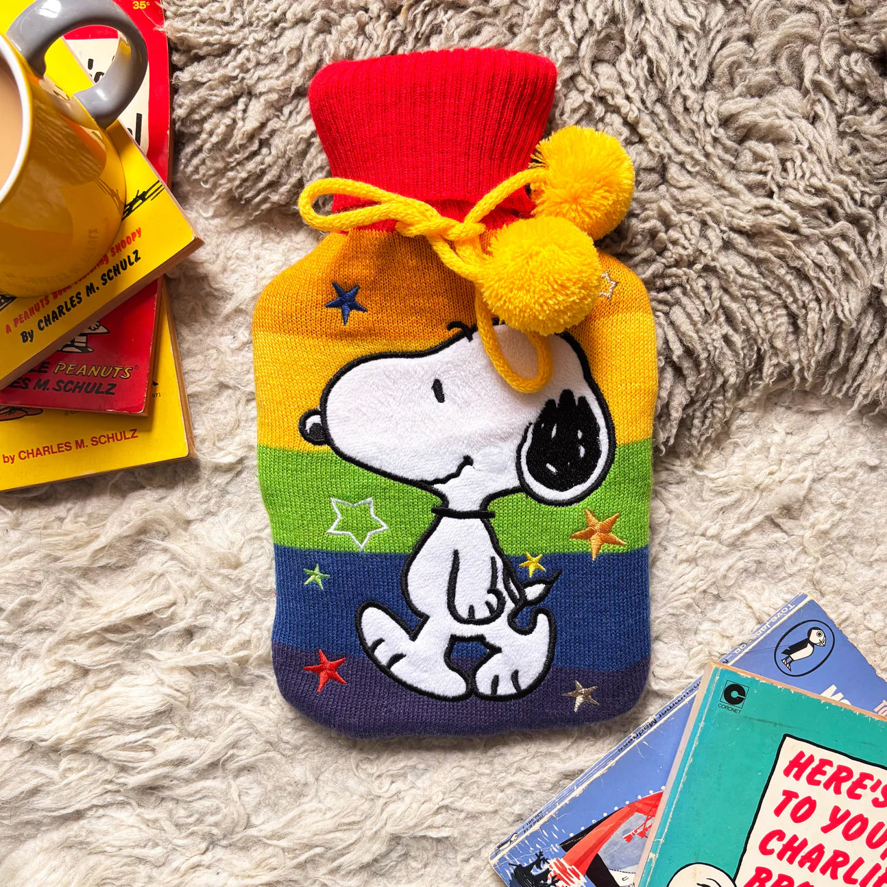 Peanuts Snoopy Rainbow Hot Water Bottle with rainbow knit cover and yellow pompoms on a fluffy rug.
