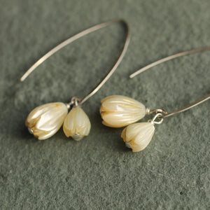 Side view of elegant drop earrings with carved mother of pearl snowdrops on curved silver wires, laid on a sage green surface.