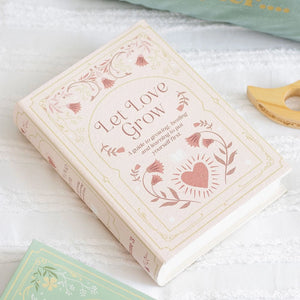 Pink book-shaped storage box with floral cover reading “Let Love Grow” on a white fabric background.