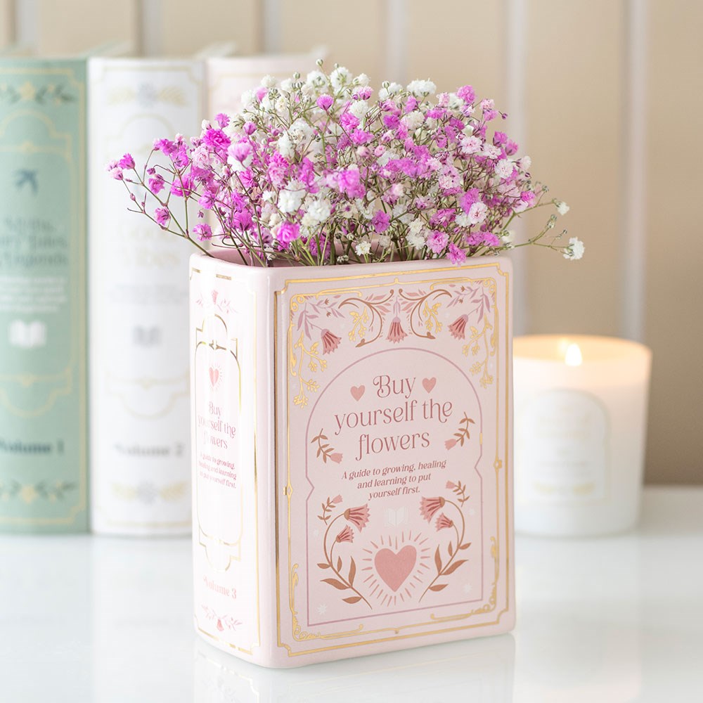 Pink ceramic book shaped vase filled with pink and white flowers displayed on a shelf beside books and a lit candle – decorative literary home décor.
