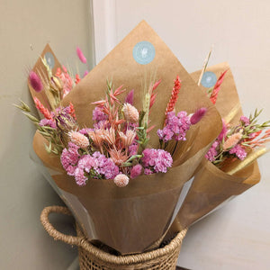 A pink dried flower arrangement wrapped in kraft paper, displayed in a woven basket.