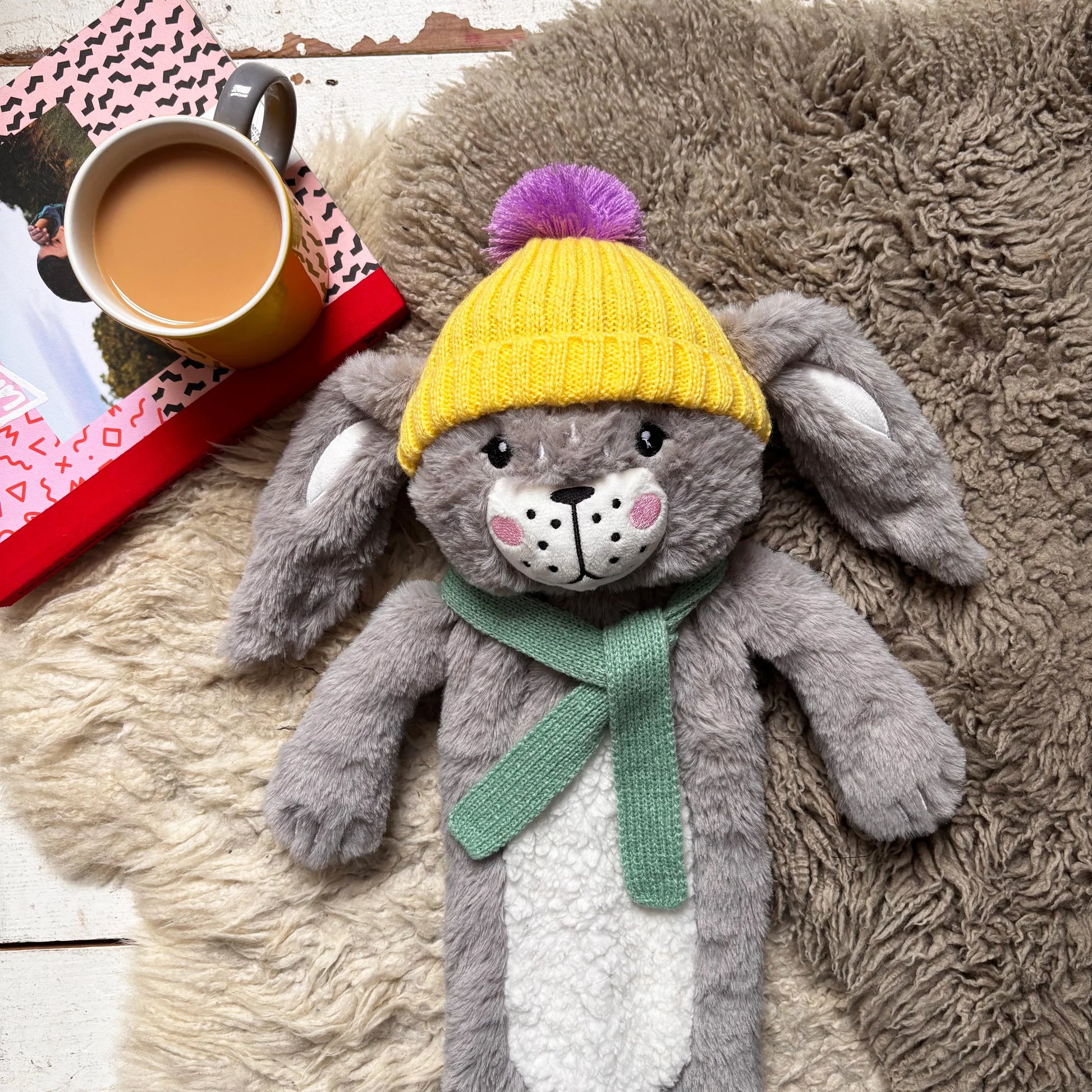 Fluffy rabbit hot water bottle lying on rug beside a mug of tea.