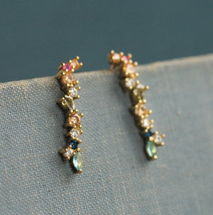 Close-up of mismatched rainbow earrings displayed vertically on textured fabric, showing gold-plated setting and multicoloured gemstones.