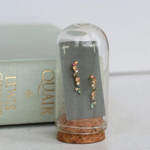 Rainbow earrings presented upright under a glass dome with cork base, styled next to a vintage book for a whimsical, curated look.