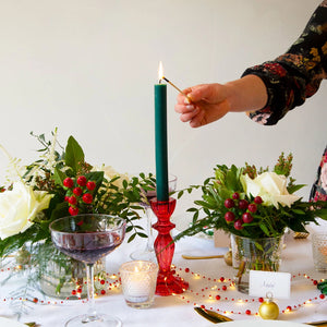 Red glass candlestick holder on a festive dinner table with green candle, white flowers, and red berry centrepieces.